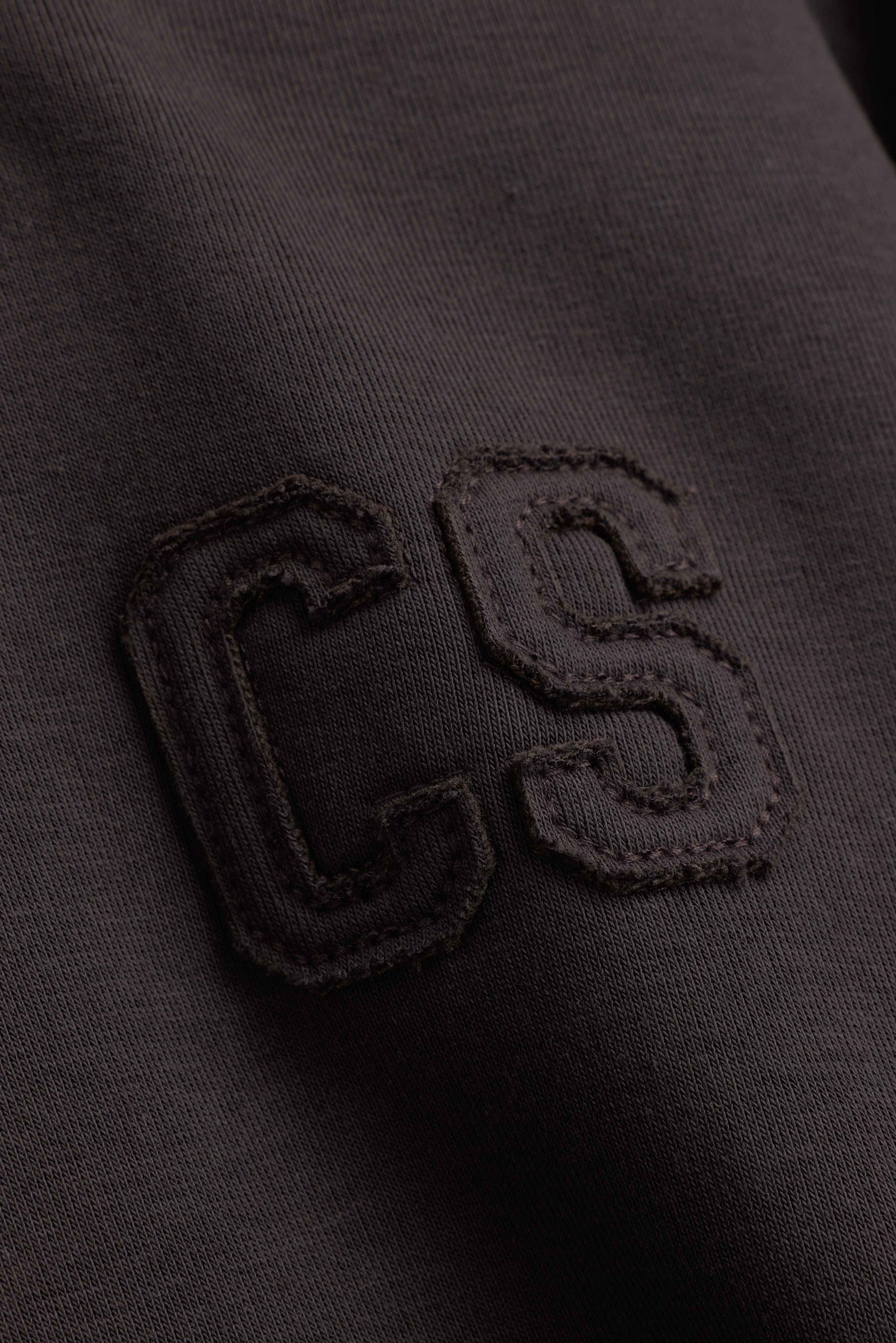 Close-up of the stitched CS logo on an espresso CASPARA CS zip hoodie.